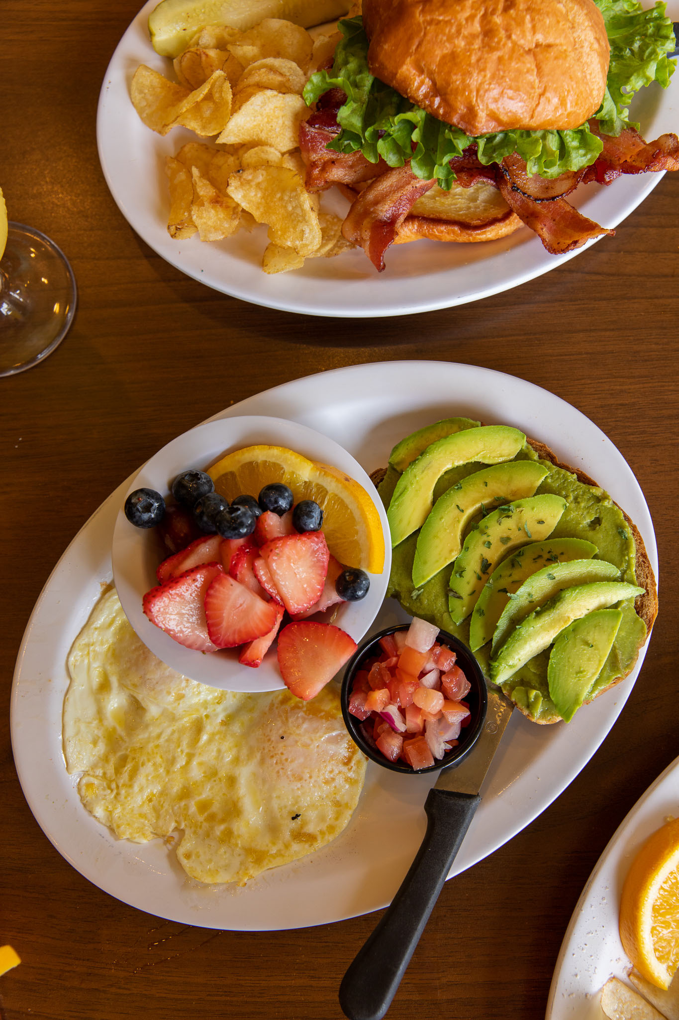 BLT sandwich fresh breakfast plate featuring avocado toast, sunny-side-up eggs, pico de gallo, and a bowl of mixed fruit