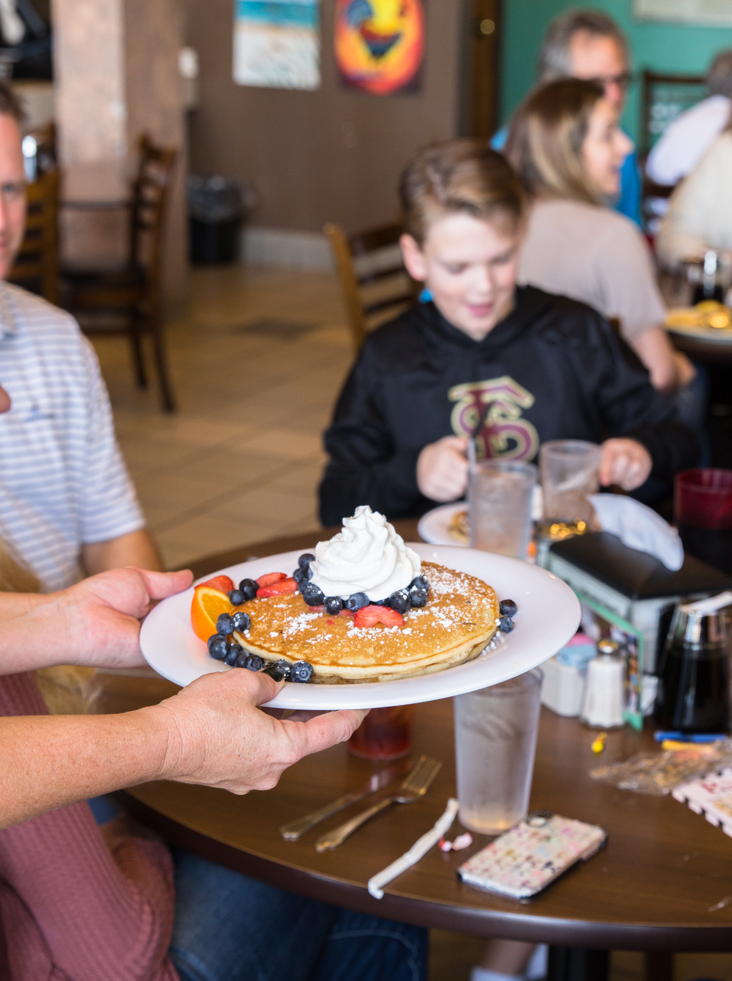 A server presents a plate of pancakes topped with blueberries, strawberries, powdered sugar, and a dollop of whipped cream.